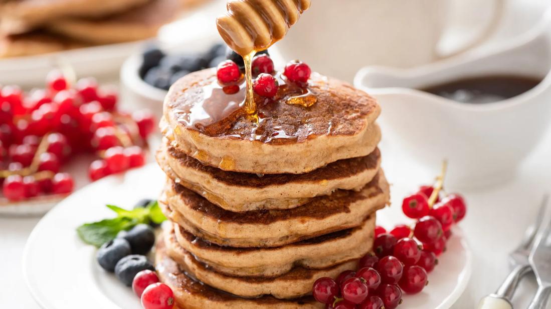 Stack of wheat and oatmeal pancakes on plate, with syrup drizzling over, with berries