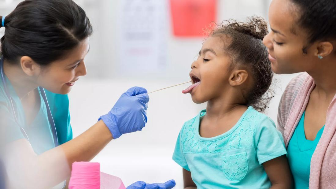 Toddler sitting on caregiver's lap receiving throat swab from healthcare provider