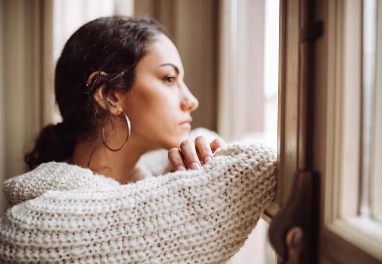 A woman with big hoop earrings and a sweater looking out a window with a pensive look on her face.