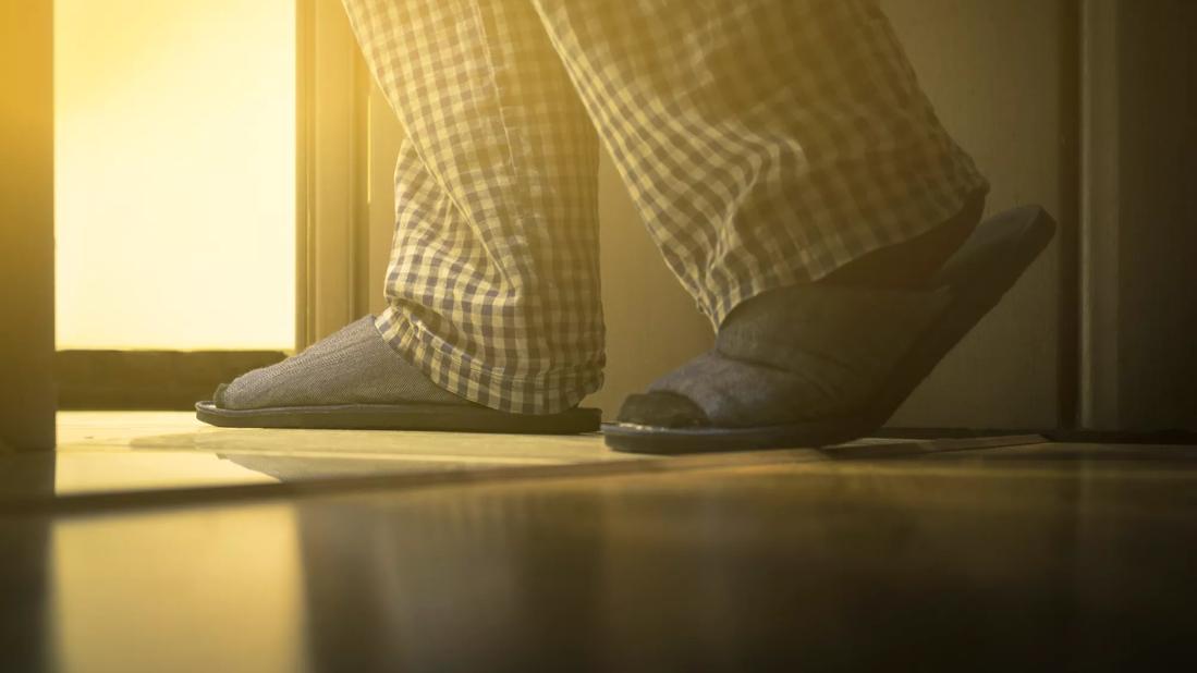 A person wearing blue plaid pajama bottoms and blue slippers walking toward the bathroom door.