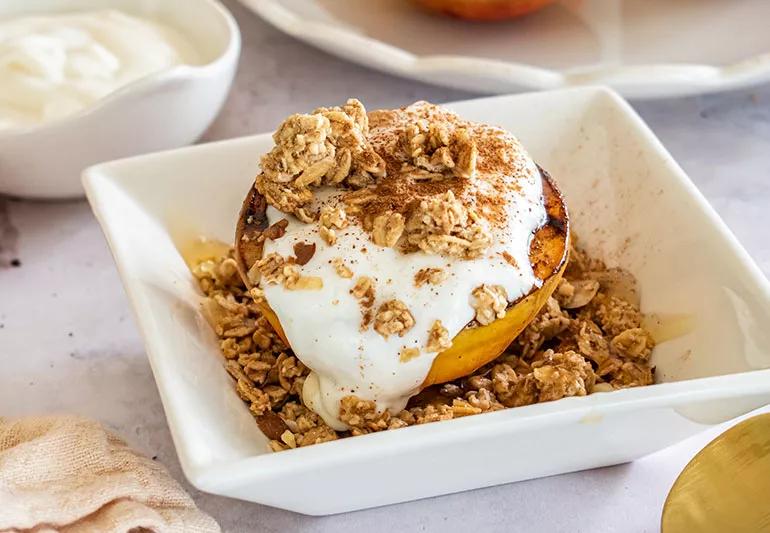 Half a grilled peach in a square white bowl topped with yogurt, cinnamon and granola