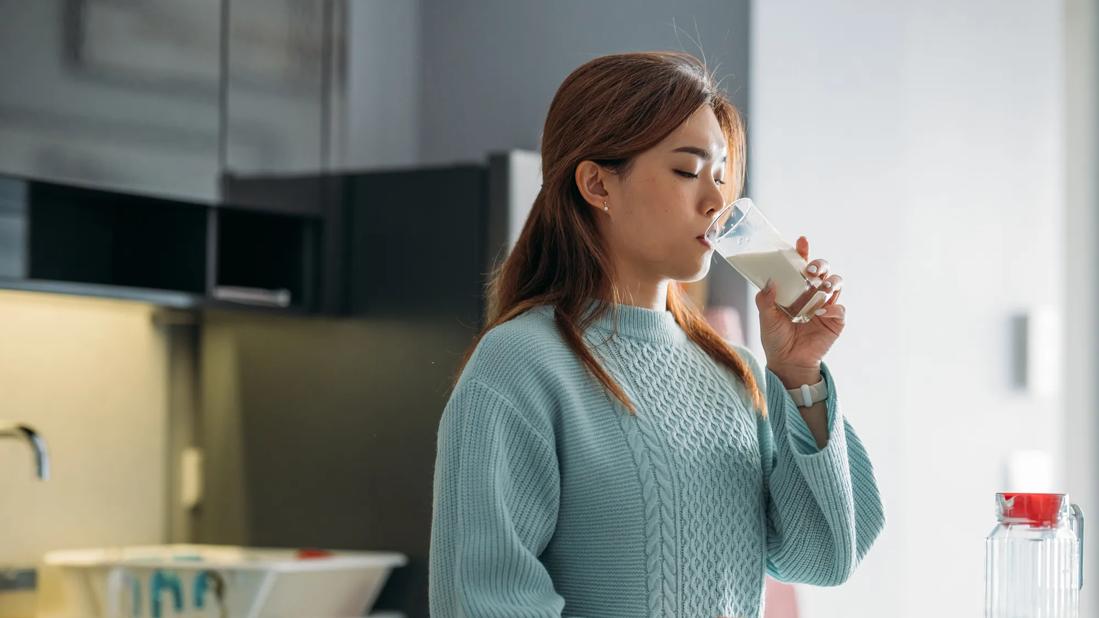 Person drinking glass of dairy product in kitchen, holding muffin in other hand