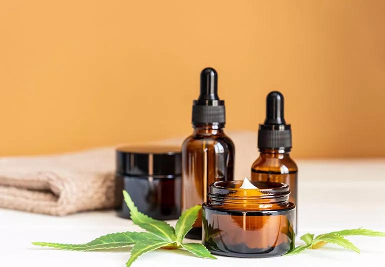 Two bottles of CBD oil and two canisters of CBD cream sit on a white counter next to marijuana leaves.