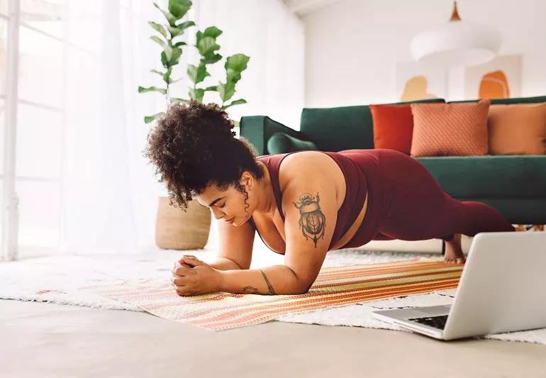 Person performing the plank position while exercising at home in living room with laptop open on floor next to them.