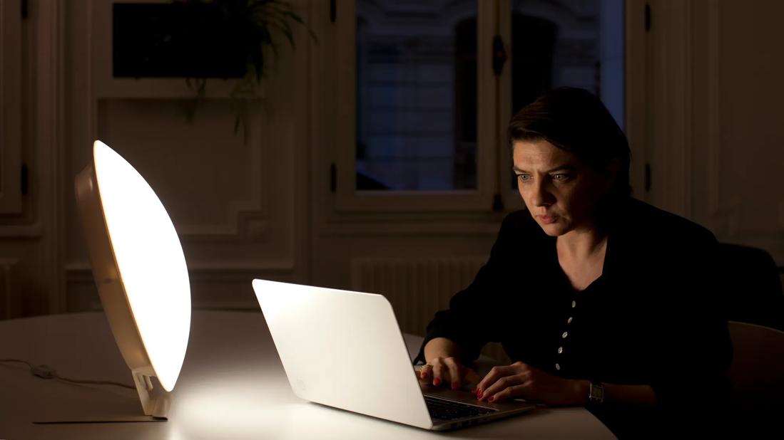 Person sitting at home desk working on laptop, with a light box for light therapy nearby