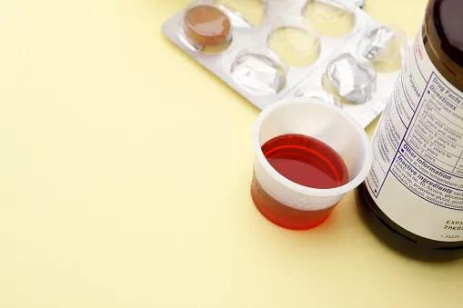 Medicine cup filled with cough syrup next to cough medicine and nearly finished packet of lozenges