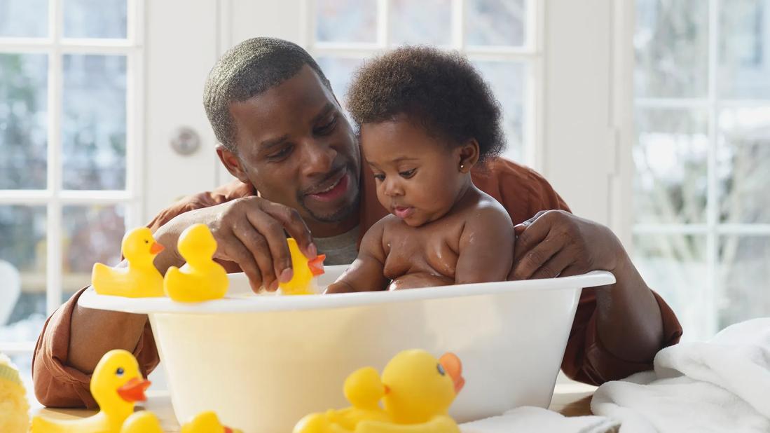 Caregiver giving young toddler a bath in plastic baby tub, with rubber duckies all around