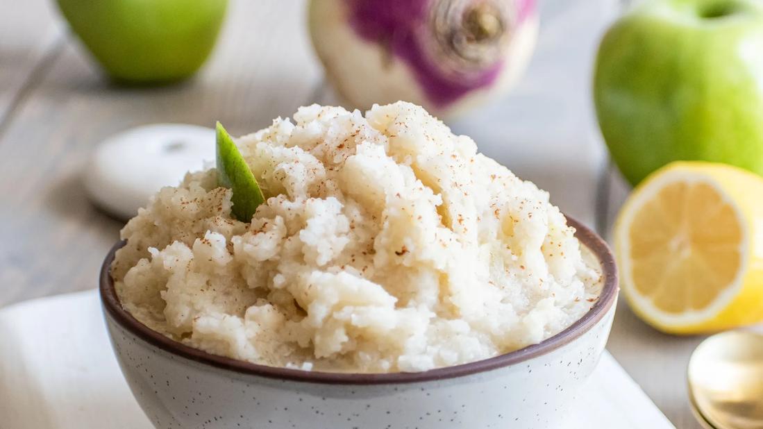 A bowl of cooked mashed turnips and Granny Smith apples, with lemons, apples and turnips in background