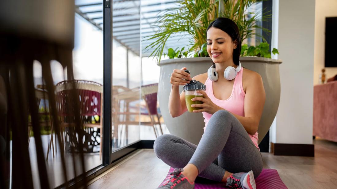 A person in exercise clothes sits on a yoga mat and holds a smoothie cup