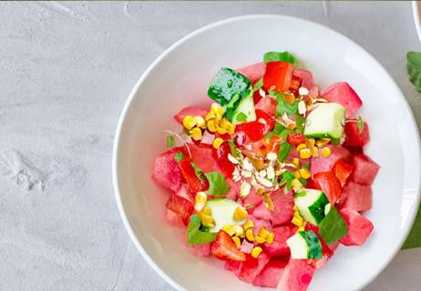 A salad in a white bowl containing watermelon, tomatoes, corn, cucumbers and almonds