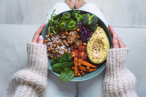 Person holding bowl of vegetarian foods on their lap, like avocado, rice, veggies and chickpeas