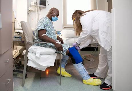 A healthcare provider checks on a patient's knee while they sit on a chair in their hospital room.