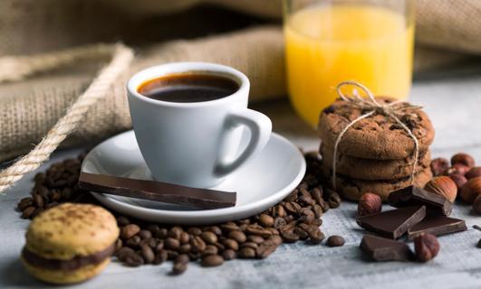 Cup of coffee on saucer, with scattered coffee beans and chocolate chunks, and baked goods