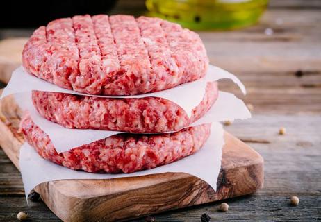 Raw hamburger patties separated by deli paper sitting on wooden cutting board.