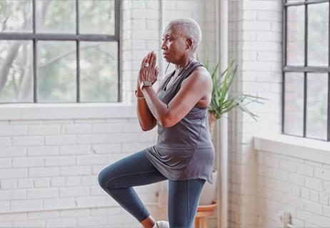 A person stands in a brightly lit room and performs a one-legged, standing yoga pose.