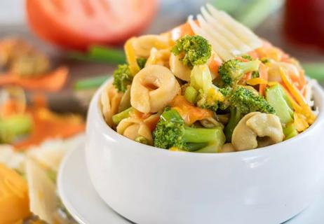 A closeup of tortellini to go in a white salad bowl with vegetables in the background.