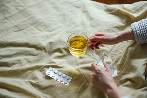 Hand holding hot glass mug of tea, with medicine packet open on bed cover