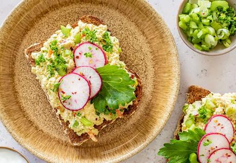 Egg salad topped with sliced radishes and cilantro sits open-faced on a piece of toast beside a small bowl of chopped celery.