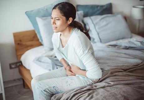 A person sitting on the edge of a bed holding their stomach in pain