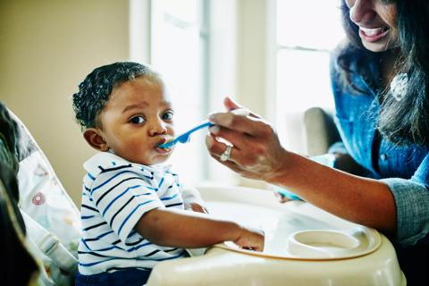 A toddler in a highchair wearing a striped shirt eats food off a spoon held by a parent