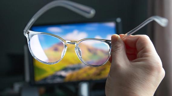 Hand holding up blue light glasses, with computer screen in background