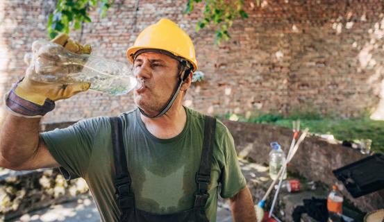 Sweaty construction worker in yellow hard hat drinking water from a plastic bottle