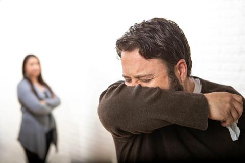 Person sneezing into arm, holding tissue, blurry person in background