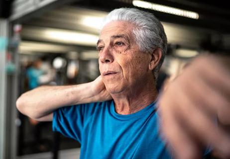 An older person holds the back of their neck while standing in a gym.