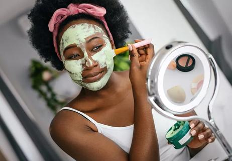A person applies a creamy green mask to their face using a brush while looking in the mirror.