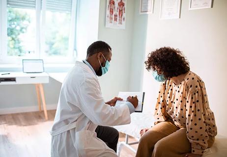 A physician wearing a face mask talking to a patient wearing a face mask in a medical office.