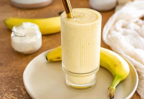 A large glass of a banana smoothie sits on a white plate next to bananas.