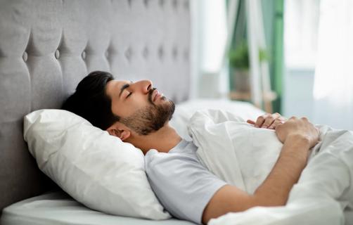 Person lying on back, hands folded against chest, sleeping in bright daylit room