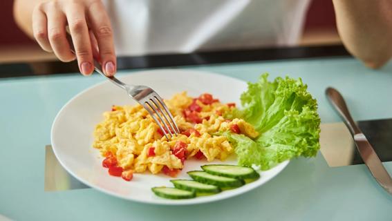 Hand holding fork, with scrambled eggs and red peppers on plate, with cucumber slices