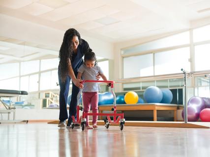 Healthcare provider helping child walk with a walker in therapy room