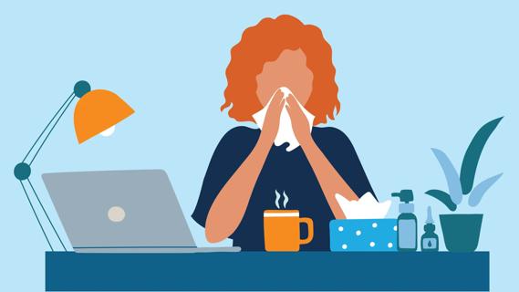 Woman blowing her nose at work desk, with hot beverage, tissues, nasal spray nearby