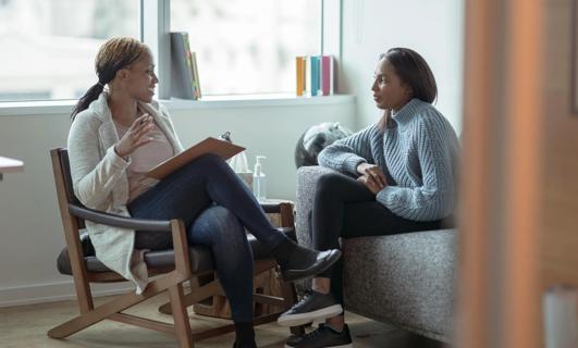 A therapist sitting in chair holding a clipboard and a patient sitting on couch talking in office