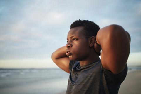 Male standing on beach with hands behind his head, staring into distance and exhaling