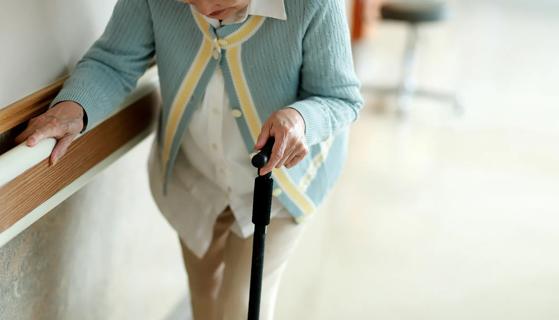 Elderly woman using a cane and handrail on wall walking down a hall