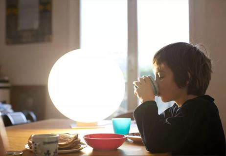 A child sits at the kitchen table eating while a large sun lamp shines natural light next to them.