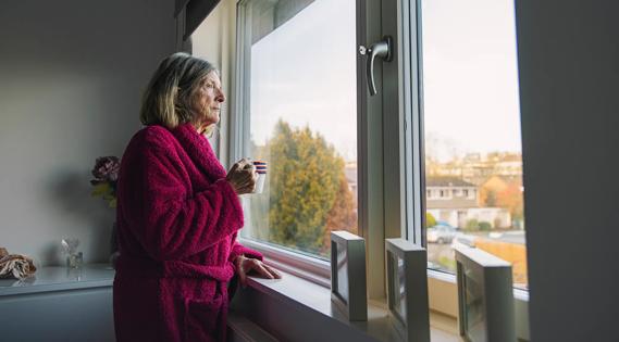 Older woman in bathrobe, holding coffee cup, looking window into the neighborhood