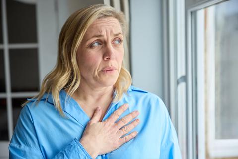 Person with furrowed brow, standing by window with their hand on their chest, taking a breath