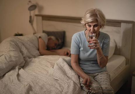 A person sitting up in bed and drinking a glass of water while another person sleeps next to them