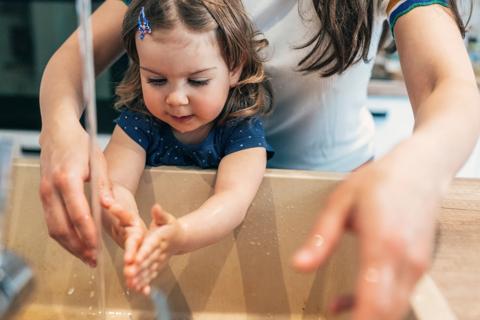Caregiver standing behind toddler at sink, helping them wash their hands