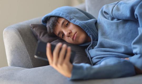 Teenage boy lying on sofa, looking through smartphone