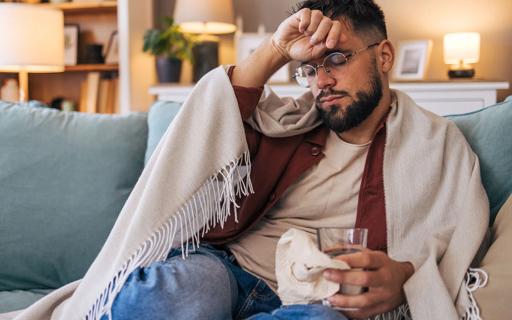 Person sick sitting on couch under throw blanket, holding glass of water and tissues, with hand to their head