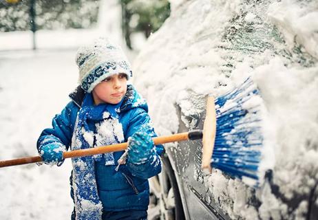 A child wearing a coat, gloves, scarf and hat sweeps snow off of a car with a broom.