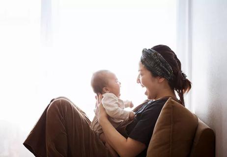 Seated mother holds small baby facing toward her as they smile at one another