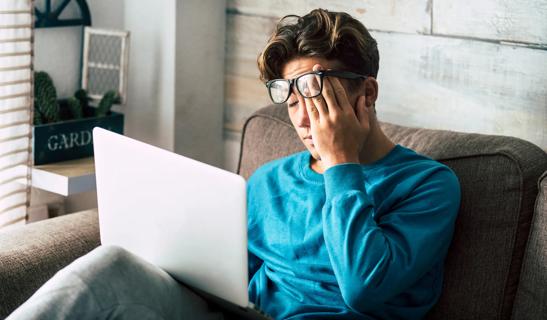 Person wearing glasses sitting on couch holding laptop, rubbing eyes, looking tired