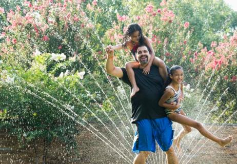 Mixed Race father holding daughters in backyard sprinkler.