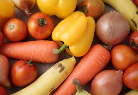 Carrots, bell peppers, onions, shallots, tomatoes and bananas are piled on a table.